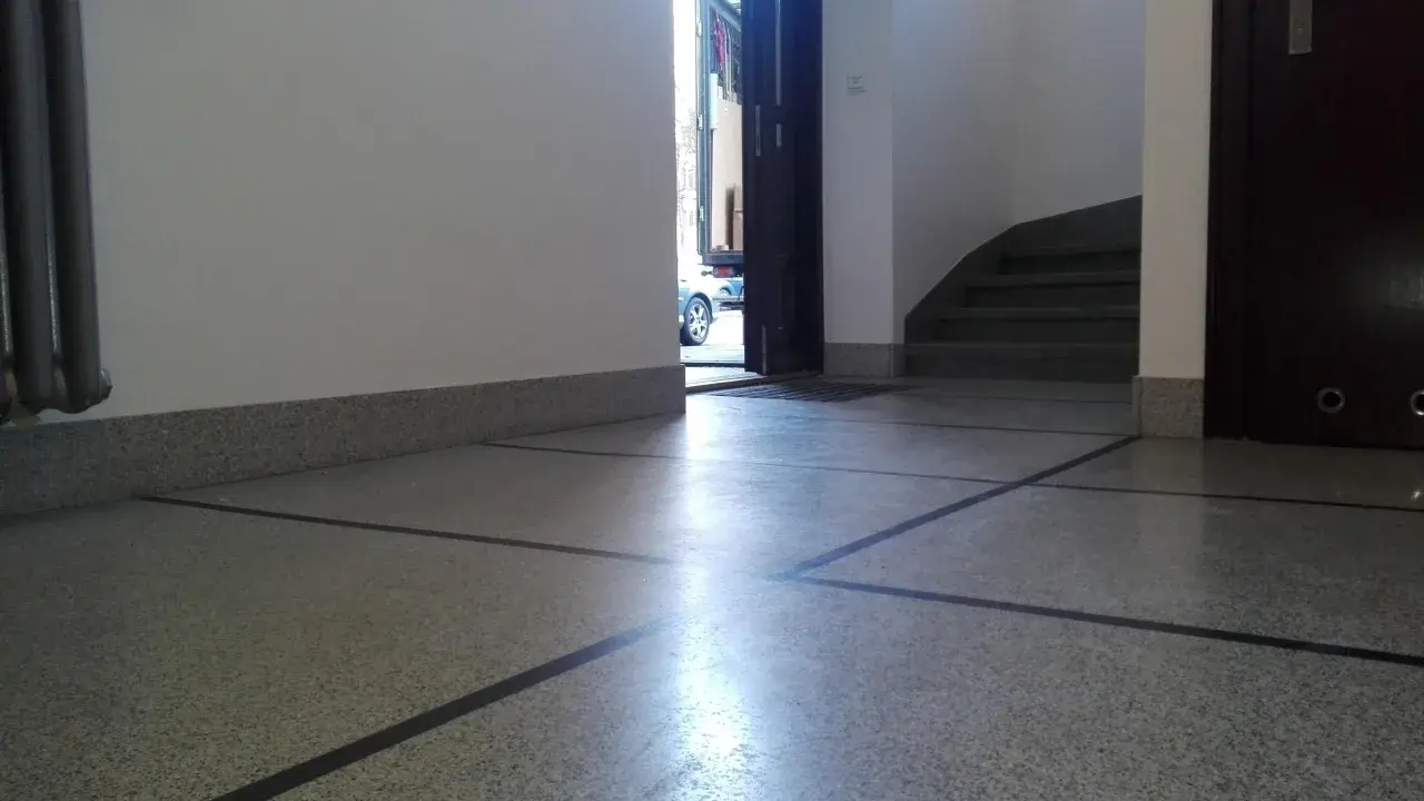 Błyszczące lastryko na podłodze klatki schodowej, z widocznymi schodami i otwartymi drzwiami na zewnątrz.