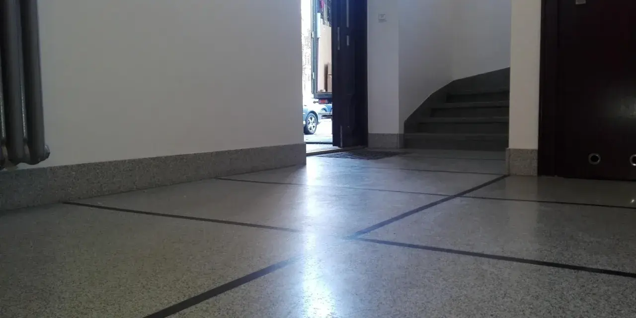 Błyszczące lastryko na podłodze klatki schodowej, z widocznymi schodami i otwartymi drzwiami na zewnątrz.