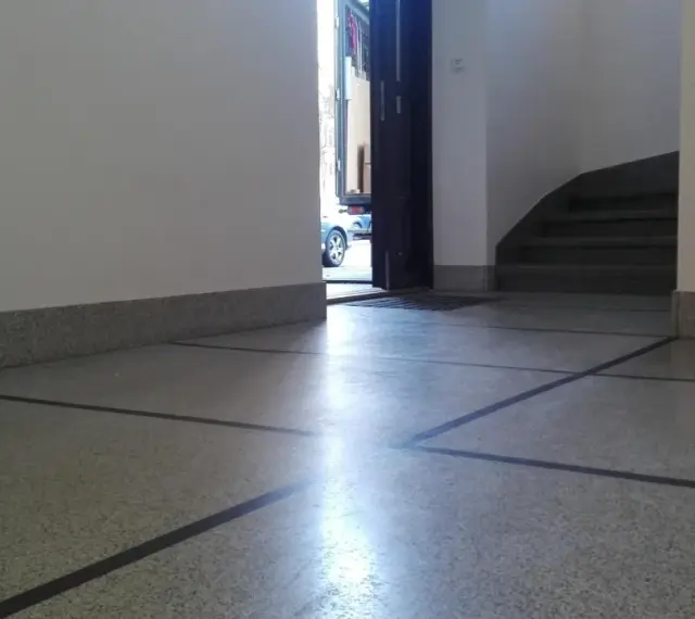 Błyszczące lastryko na podłodze klatki schodowej, z widocznymi schodami i otwartymi drzwiami na zewnątrz.