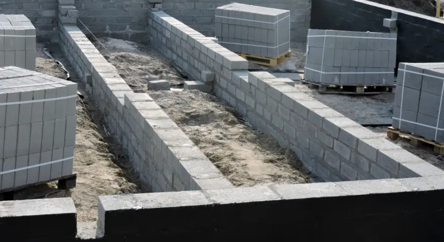 Fundament budowy z bloczków betonowych, przygotowany pod izolację przeciwwilgociową. Materiały budowlane czekają na użycie.